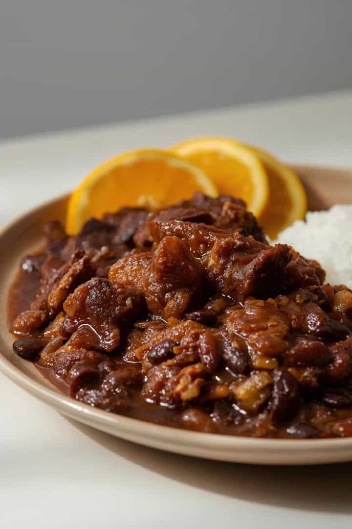 Low side-angle food photography showing feijoada, black beans and meats layered thickly, not watery. Plate with rice and oranges visible at the edge. Clean composite styling. Neutral daylight. Ver